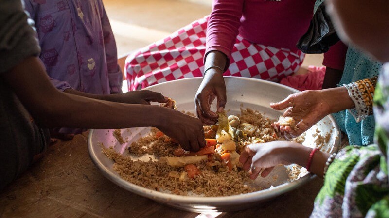 WFP/Jean-Baptiste Joire Several hands reach into a large communal metal platter of rice and vegetables, including carrots and what appears to be cassava or yam, eating with their fingers in the traditional manner. WFP/Jean-Baptiste Joire